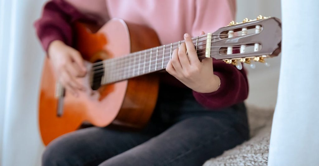 Faceless skilled female musician playing acoustic guitar while sitting on comfortable bed near white curtains during music performance at home